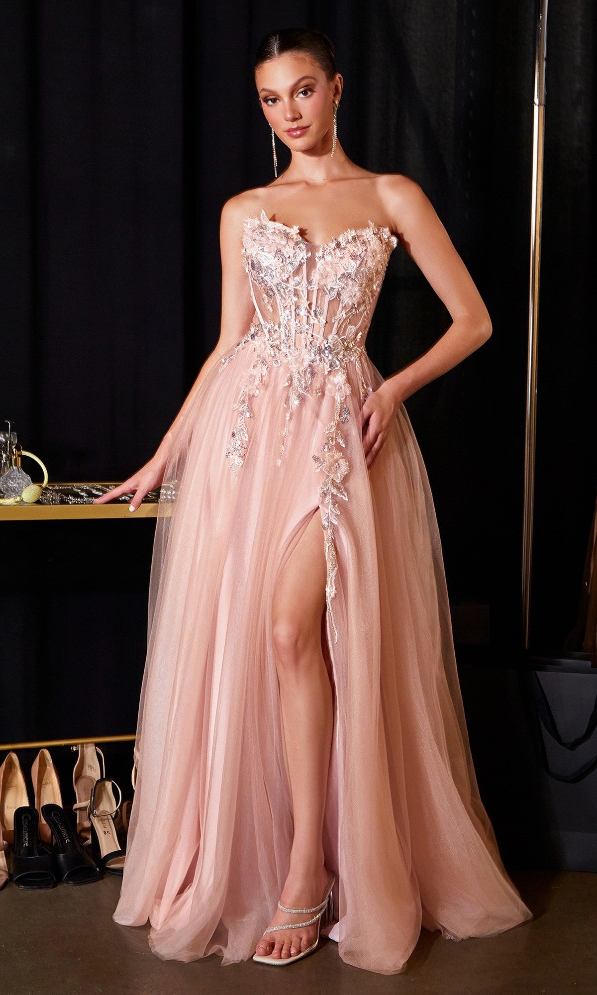 A woman stands indoors wearing the Ladivine CD2426, a strapless, light pink long prom dress with floral embroidery and a high slit, posing beside a table with jewelry and shoes.